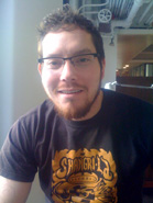 A man with short brown hair, glasses, and a goatee wearing a black t-shirt with a gold graphic.