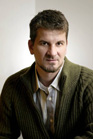 A man with short brown hair and a goatee wearing a white shirt and a green ribbed cardigan.