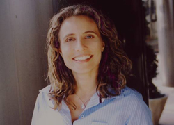 A smiling woman with wavy brown hair wearing a light blue button-down shirt.