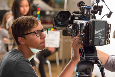 A person with glasses looking through the viewfinder of a large professional video camera on a tripod.