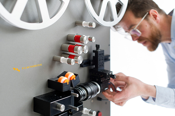 A man with glasses leans in to adjust a black lens and mechanical parts on a grey film projector.