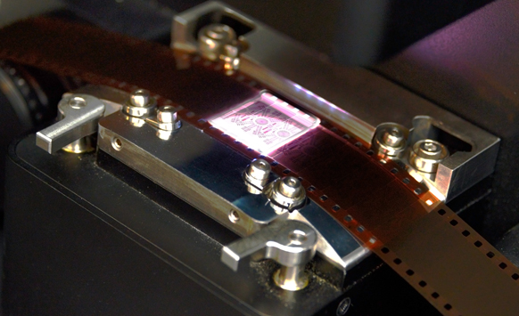 A strip of brown film passing through a metal mechanical device with bright light shining through a frame.