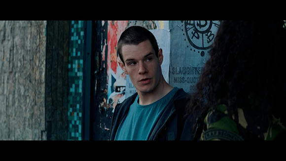A young man with short hair looks toward a person with dark curly hair in front of a graffiti wall.