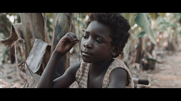 A young child with dark curly hair looking thoughtfully to the side in a tropical outdoor setting.