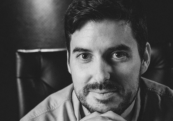 Close-up black and white portrait of a man with dark hair and a beard looking directly at the camera.