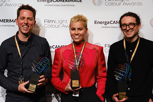 Three smiling people standing together holding colorful wire-sculpture trophies against a branded backdrop.
