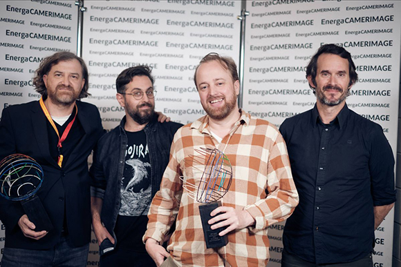 Four men stand side-by-side in front of a branded backdrop, two holding awards.