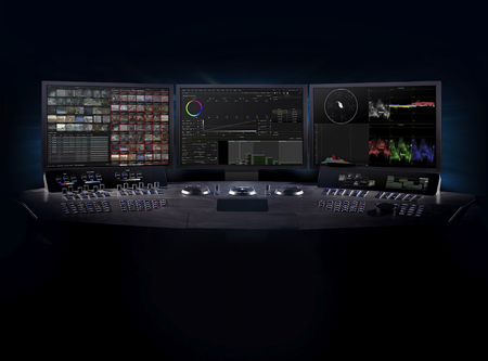 Three computer monitors displaying video editing software above a large control panel with various knobs and sliders.