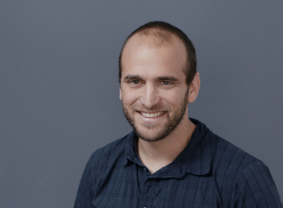A smiling man with short dark hair and a beard wearing a dark blue checkered button-down shirt.
