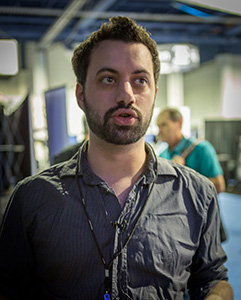A man with dark hair and a beard wearing a grey button-down shirt speaks in a studio.