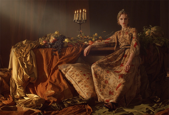 A woman in a long patterned dress sits by a table overflowing with fruit and lit candles in a dimly lit room.