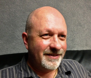 A bald man with a grey goatee and a striped shirt smiling against a grey background.