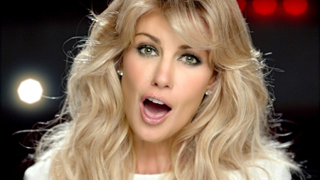A blonde woman with green eyes singing with her mouth open against a dark, blurred background.