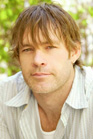 A man with shaggy light brown hair and a light-colored striped shirt looking forward.