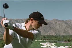 A person in a black cap and white shirt swings a golf club on a course with mountains in the background.