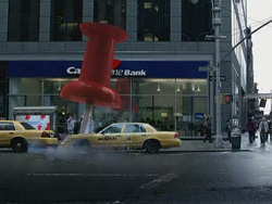 A massive red pushpin sculpture stands on a city street in front of a bank building with yellow taxis passing by.