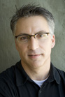 A man with short graying hair and glasses wearing a black polo shirt against a gray wall.