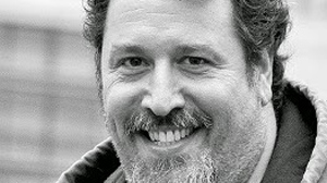 Black and white close-up portrait of a man with wavy hair and a beard, smiling at the camera.