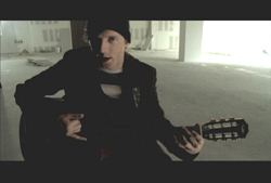 A man in a dark jacket and beanie plays an acoustic guitar in a bright, empty room.