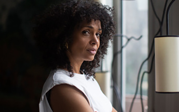 A woman with dark curly hair and a white sleeveless top looks toward the camera in a dimly lit room.