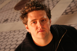 A man with spiked brown hair wearing a black jacket, looking forward against a patterned background.