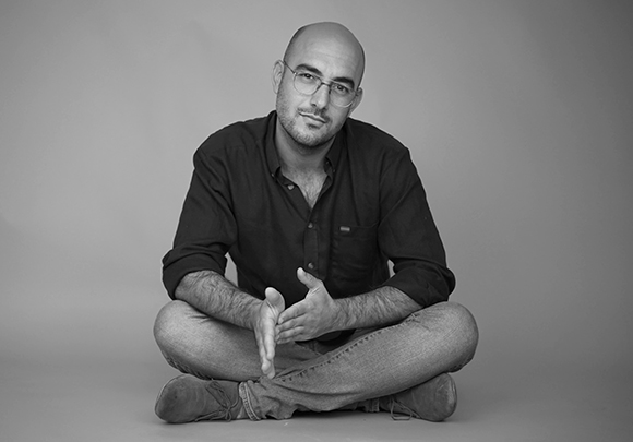 A bald man wearing glasses and a dark button-down shirt sits cross-legged against a plain background.