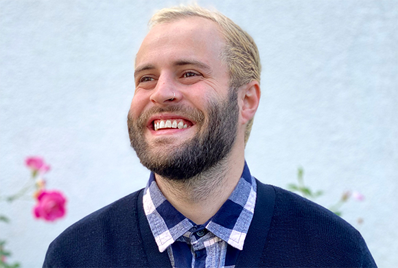 A smiling man with blonde hair and a dark beard wearing a blue checkered shirt and dark cardigan.
