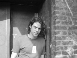 A man with wavy hair leans against a brick wall in a black and white portrait.