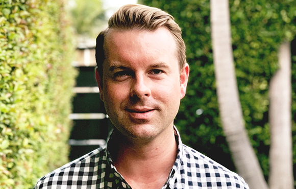 A man with short blonde hair wearing a black and white checkered shirt, smiling in front of green foliage.