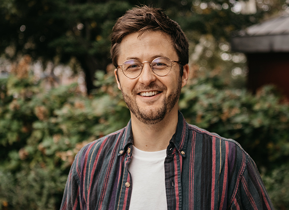A smiling man with short brown hair, glasses, and a beard wearing a striped button-down shirt.