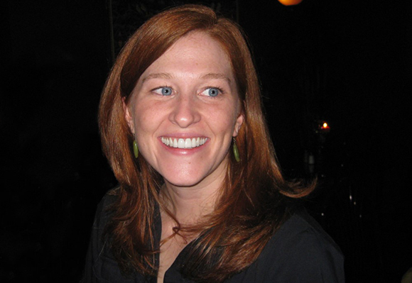 A smiling woman with long reddish-brown hair and green hoop earrings.