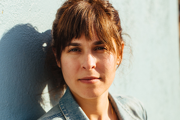 A woman with brown hair and bangs looks directly at the camera while leaning against a light blue wall.
