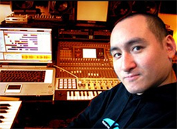 A man smiling in a music studio filled with mixing consoles and computer monitors.