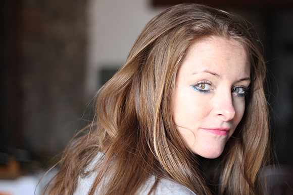 A woman with long brown hair and blue eyeliner looks toward the camera over her shoulder.