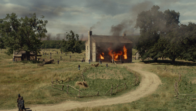 A stone house engulfed in flames in a grassy field with smoke rising into a cloudy sky.