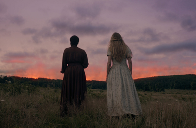 Two women in long dresses stand in a grassy field facing a sunset with purple and orange clouds.