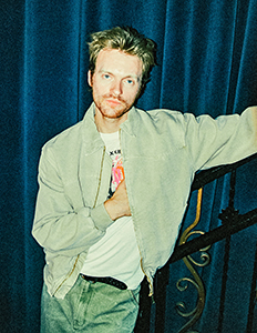A man with blond hair wearing a light jacket and white t-shirt leans against a railing in front of blue curtains.