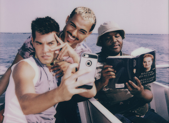 Three men on a boat; one takes a selfie while another reads a book titled Madam Secretary.