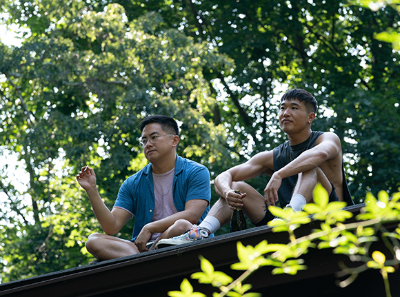 Two men sit on a dark roof edge against a backdrop of lush green trees.
