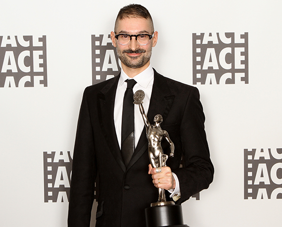 A man with glasses and a mustache wearing a black suit and tie, holding a silver trophy against a white backdrop.