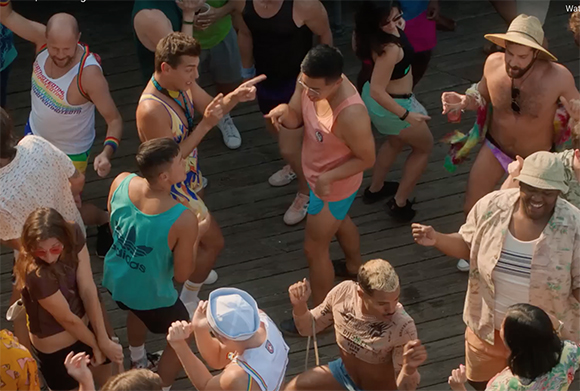 A diverse group of people in colorful summer clothing dancing together on a wooden boardwalk.