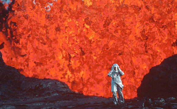 An astronaut in a silver suit stands before a massive, glowing orange and red lava eruption.