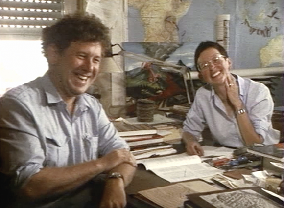 A man and a woman sit at a cluttered desk, laughing together in an office.