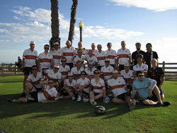 A large group of cyclists in matching white jerseys posing together on a grassy field under a blue sky.