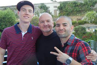 Three men posing together outdoors, one in a striped polo, one in black, and one in a red plaid shirt.