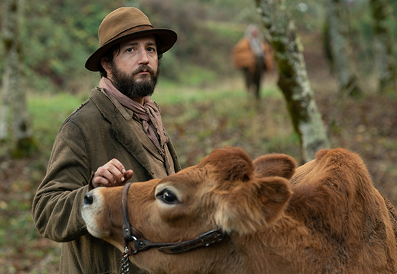A man wearing a brown hat and jacket stands in a forest next to a brown calf.
