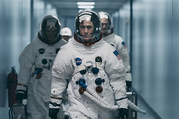 Three astronauts in white NASA spacesuits walk down a sterile, brightly lit hallway.