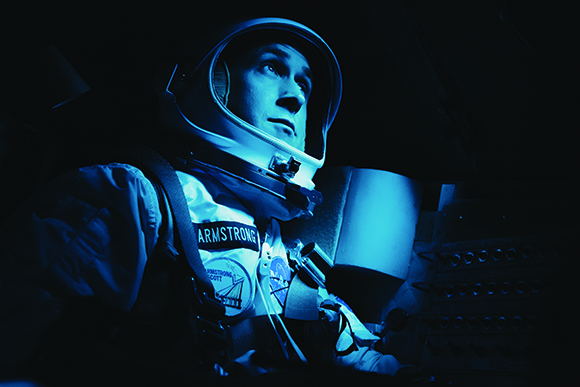 A person in a white spacesuit and helmet looking upward, lit by a cool blue light against a dark background.