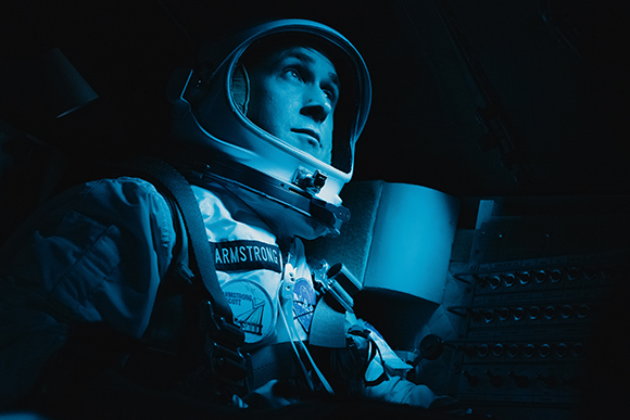 An astronaut wearing a helmet and spacesuit looks upward in a dark, blue-lit spacecraft cockpit.