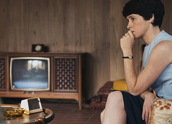 A woman with short dark hair sits with her hand to her mouth, looking toward a vintage television in a wood-paneled room.